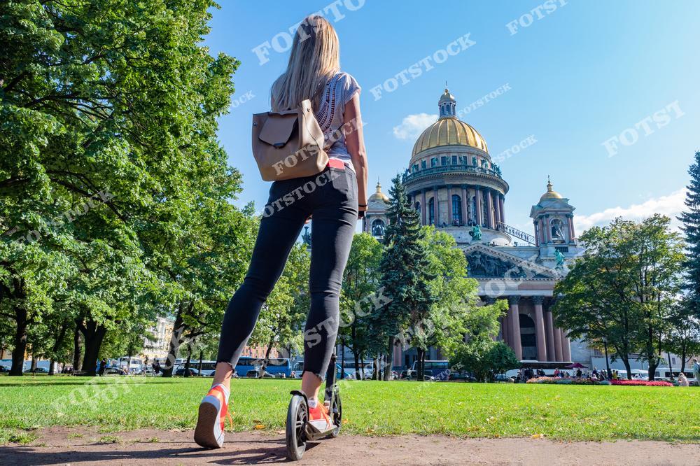 На снимке изображена женщина на самокате, остановившаяся рядом с Исаакиевским собором в Санкт-Петербурге. Величественный собор с его золотым куполом и классической архитектурой возвышается на фоне чистого голубого неба. Вокруг здания зелень летнего парка, создающая уютную атмосферу для прогулок. Исаакиевский собор является одной из знаковых достопримечательностей города, привлекающей туристов своим историческим и архитектурным значением.